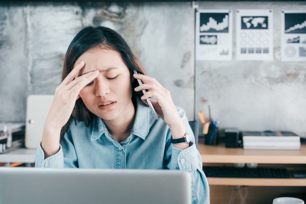 Stressed Asian creative designer woman cover her face with hand and feel upset while talk on mobile phone with customer in front of laptop computer on desk at office,Stress office lifestyle concept